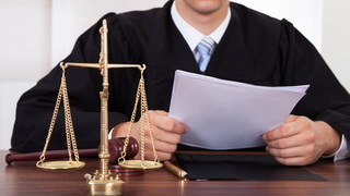Foto: Richter sitzt mit Papier in der Hand am Tisch mit Waage und Hammer
